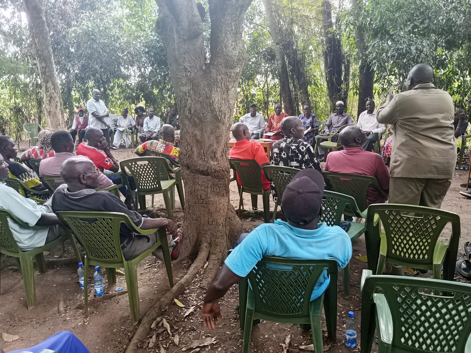 A speaker addressing community members seated under trees